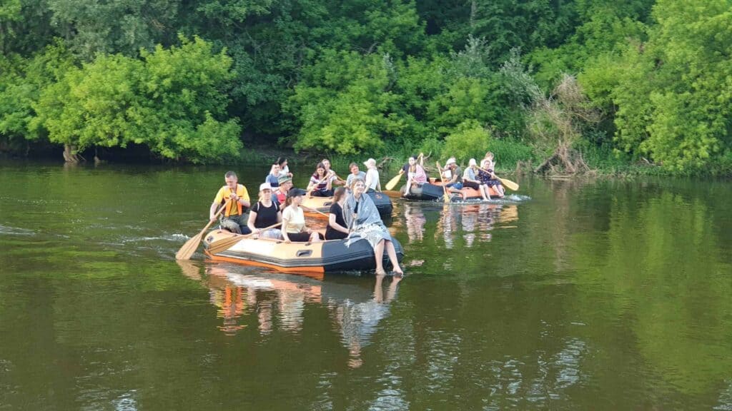 Trzy pontony z Bazy Pontonowej, każda z grupami ludzi, płyną łagodnie przez spokojną rzekę, otoczoną gęstym zielonym listowiem. Spokojna atmosfera doskonale odzwierciedla przygodę oferowaną przez EVERAL w tym naturalnym sanktuarium.