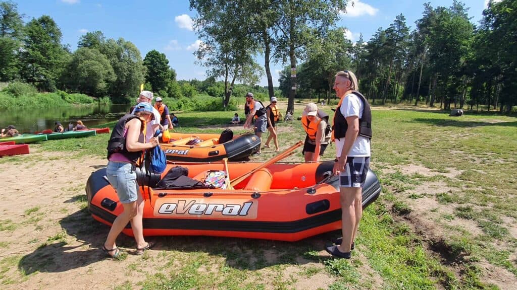Na bazarze pontonowym ludzie przygotowują pontony nad brzegiem rzeki w słoneczny dzień, ubrani w kamizelki ratunkowe i codzienne, letnie ubrania.