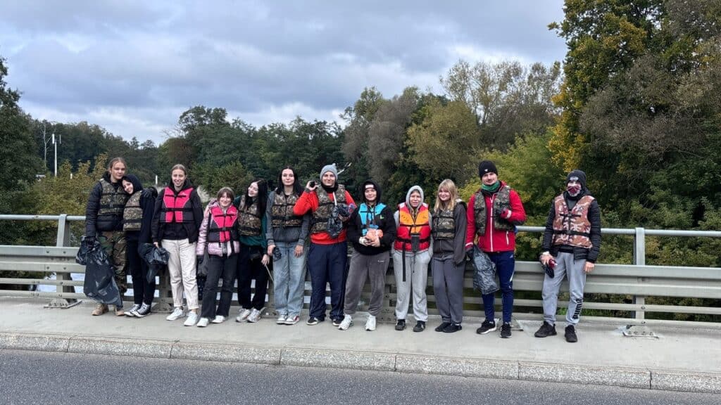 Grupa trzynastu osób w kamizelkach ratunkowych stoi na moście, pozując do zdjęcia podczas akcji Czyste Rzeki Jesiennej, z drzewami i zachmurzonym niebem w tle.