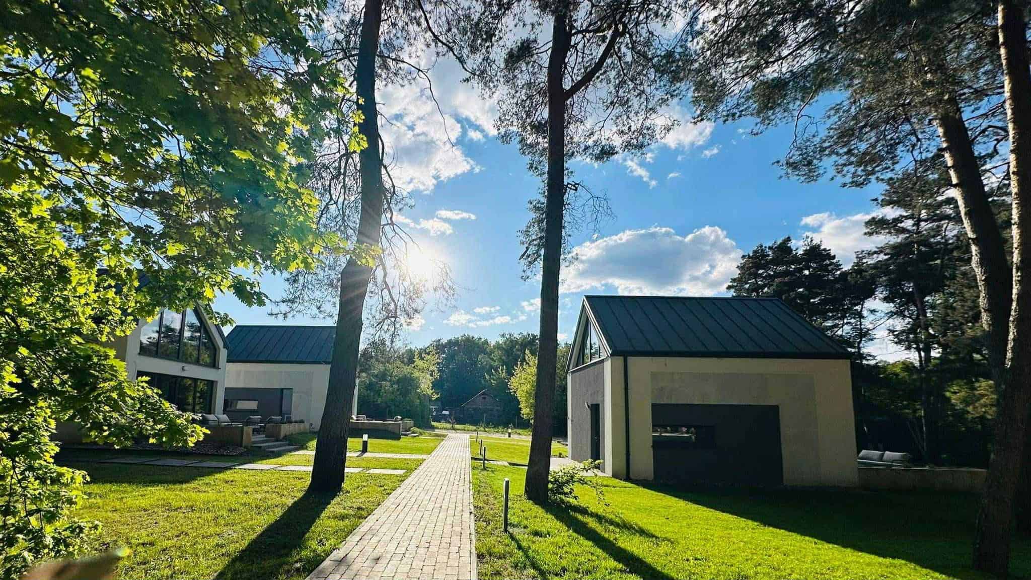 Wybrukowany chodnik biegnie między nowoczesnymi białymi domami z ciemnymi dachami w OŚRODKU WYPOCZYNKOWYM DWA ŚWIATY, otoczony drzewami i trawą pod jasnoniebieskim niebem z rozproszonymi chmurami.