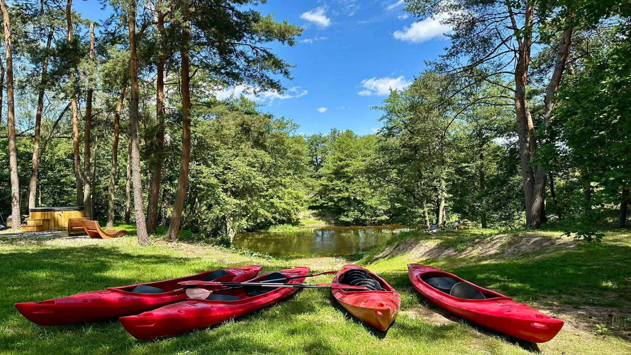 Pięć czerwonych kajaków i wioseł spoczywa na trawie w pobliżu małego stawu w ośrodku wypoczynkowym Dwa Światy, otoczonym wysokimi drzewami i zielenią pod częściowo zachmurzonym niebem.